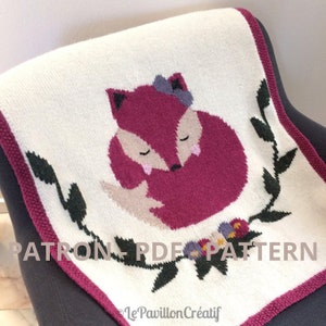 May include: A knitted baby blanket with a pink fox design. The fox is surrounded by green leaves and flowers. The blanket has a white background with a pink border. The text "PATRON - PDF - PATTERN" is printed on the blanket.