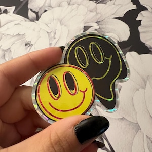 May include: Two holographic stickers featuring smiley faces. One is a classic yellow smiley face with a red outline. The other is a black, distorted smiley face. The stickers are held against a floral background.