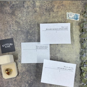May include: A gold ring with a teardrop-shaped stone in a beige box, with three white cards asking questions like "Where did you meet the newlyweds?" and "Share your favorite memory from today."