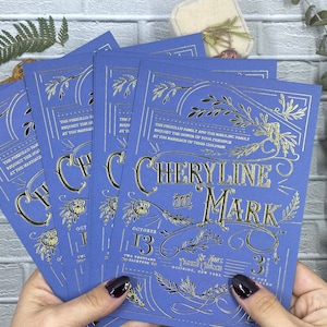May include: Four blue wedding invitations with gold foil lettering. The invitations feature the names "Cheryline and Mark", the date "October 13", and event details. The invitations are held in front of a white brick wall.