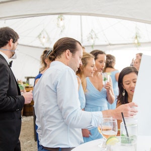 May include: A group of people stand around a table with drinks and a white board. The people are wearing casual clothing and smiling. The board is blank and has a black easel.
