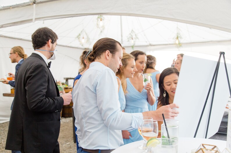 May include: A group of people stand around a table with drinks and a white board. The people are wearing casual clothing and smiling. The board is blank and has a black easel.