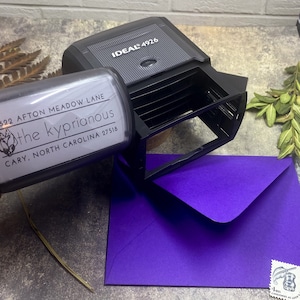 May include: A black Ideal 4926 address stamp with a clear address label, next to a purple envelope. The address label reads "622 Afton Meadow Lane, the kyprianous, Cary, North Carolina 27518". A US postage stamp is visible.
