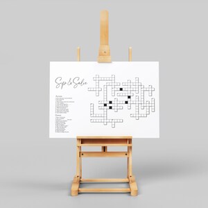 May include: A white crossword puzzle on an easel with the title "Sip & Solve". The puzzle has a grid of squares with black squares for clues. The puzzle is on a white background.