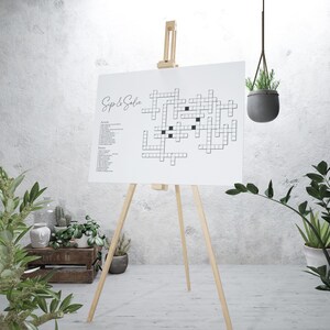 May include: A white crossword puzzle on an easel with the title "Sip & Solve". The puzzle is set up with black squares for the clues and white squares for the answers. The puzzle is on a white background.