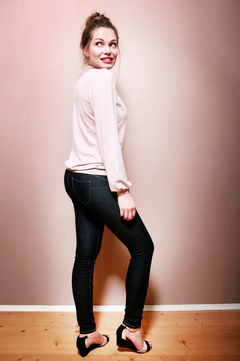 K&ouml;nnte beinhalten: Eine Frau tr&auml;gt ein hellrosa Langarmhemd, eine dunkelblaue Skinny Jeans und schwarze und goldene High Heels. Sie steht vor einer hellrosa Wand.