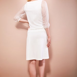 May include: A white dress with a scoop neck and sheer dotted sleeves. The dress has a skirt that falls to the knee. The model is wearing white heels.