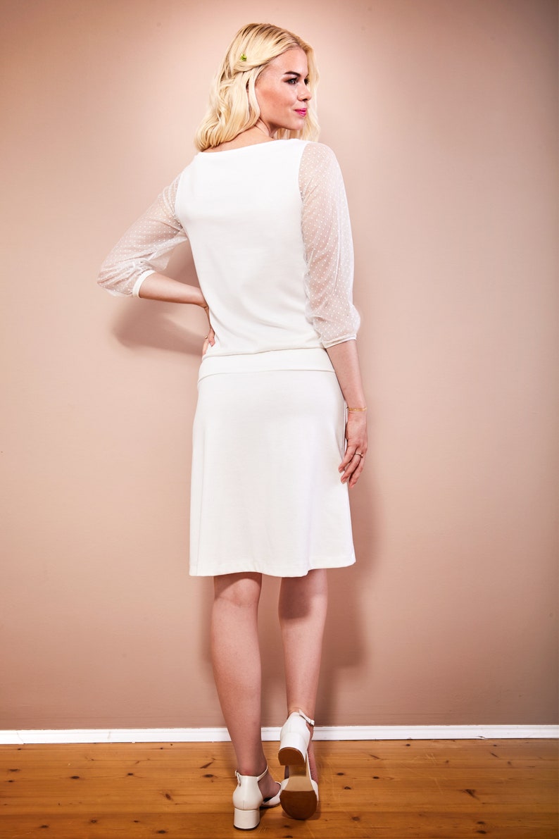 May include: A white dress with a scoop neck and sheer dotted sleeves. The dress has a skirt that falls to the knee. The model is wearing white heels.