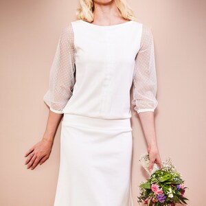 May include: A white dress with a fitted bodice and a-line skirt. The dress has sheer sleeves with polka dots. The model is holding a bouquet of flowers.