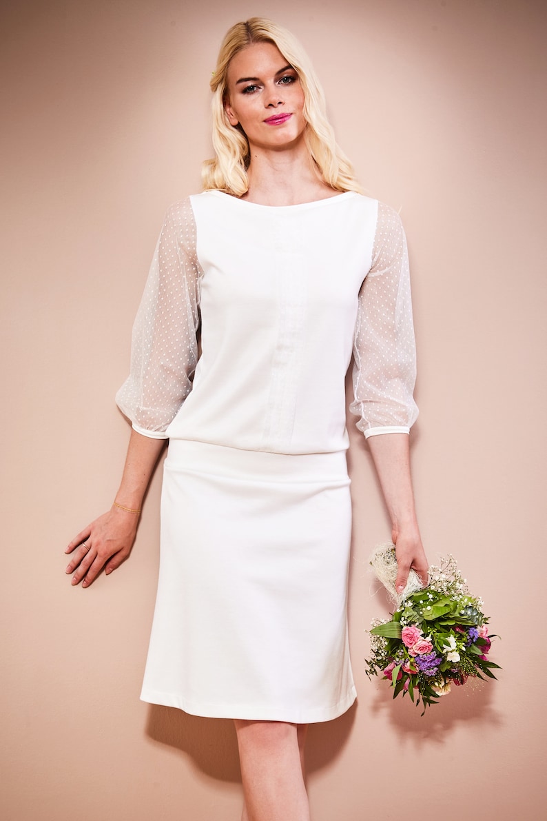 May include: A white dress with a fitted bodice and a-line skirt. The dress has sheer sleeves with polka dots. The model is holding a bouquet of flowers.