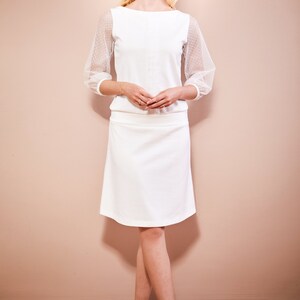May include: A white top with sheer polka dot sleeves and a white skirt. The model is wearing white shoes.