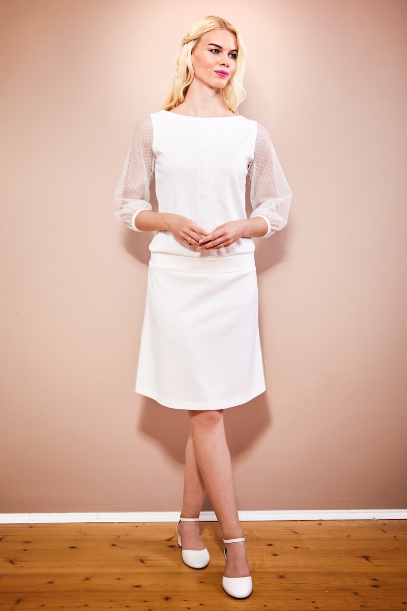 May include: A white top with sheer polka dot sleeves and a white skirt. The model is wearing white shoes.