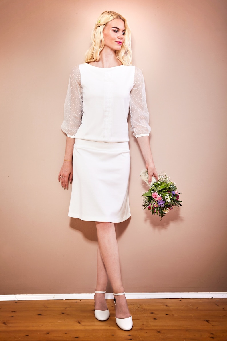 May include: A white dress with a fitted bodice and a-line skirt. The dress has sheer polka dot sleeves and a simple neckline. The model is holding a bouquet of flowers.