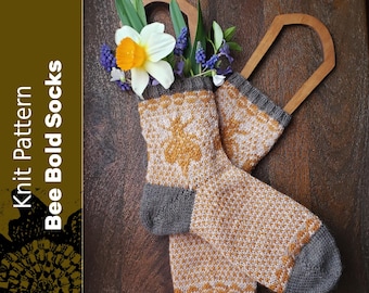 Peut inclure: Une paire de chaussettes tricotées avec un motif d'abeille dans des tons de jaune et de blanc. Les chaussettes sont présentées sur une surface en bois avec un arrangement floral.