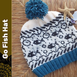 May include: A white knitted hat with a blue pom pom and a black and white fish pattern. The hat has a blue brim.