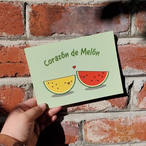Op de afbeelding: Een groene kaart met de tekst "Corazón de Melón" en twee cartoonwatermeloenen met gezichten. De watermeloen links is geel en de watermeloen rechts is rood.