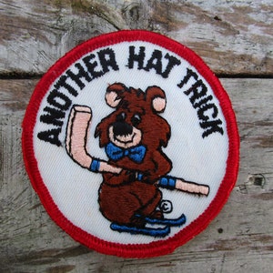 May include: Embroidered patch with a cartoon bear wearing a blue bow tie and skis, holding a hockey stick. The patch has a white background with the text "ANOTHER HAT TRICK" in a circular pattern, framed by a red border.