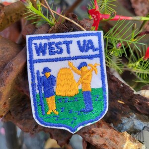 May include: Embroidered patch with a shield design featuring the text "WEST VA" in blue. The patch depicts figures in blue and yellow, a yellow haystack, and green grass. The patch is bordered in blue and white.