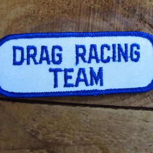 May include: A rectangular patch with a white background and blue embroidered text that reads "DRAG RACING TEAM." The patch has a blue border and is likely for a racing team.