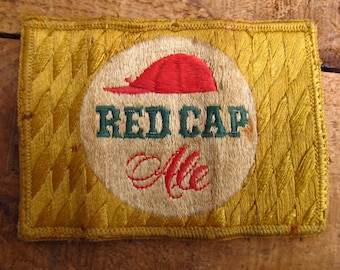 Peut inclure: Un patch brodé vintage avec une casquette rouge et les mots "RED CAP Ale" en lettres vertes et blanches sur un fond doré.