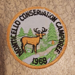 May include: A round, embroidered patch with a brown deer in a forest setting. The patch is beige with green and brown embroidery. The text "Monticello Conservation Campcraft 1958" is embroidered in black around the edge.