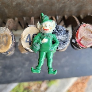May include: A green plastic figurine of a smiling elf wearing a green hat and holding a green bucket. The elf is standing on a wooden surface with a number 40 on it.