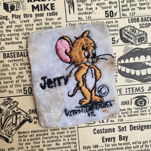 May include: A vintage embroidered patch featuring Jerry the mouse from the Tom and Jerry cartoon. The patch is white with brown and pink embroidery. The text "Jerry" is embroidered in brown below the image.