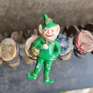 May include: A green plastic figurine of a leprechaun holding a pot of gold. The leprechaun is wearing a green hat with a gold trim and a green suit with gold buttons. The figurine is about 3 inches tall.