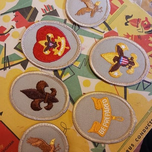 May include: A collection of ten vintage Boy Scout merit badges. The badges are circular and feature various designs, including a heart, a fleur-de-lis, an eagle, and a banner with the words "Be Prepared".