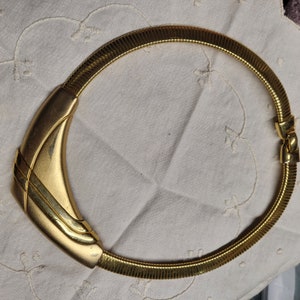 May include: A gold-toned necklace with a geometric, curved pendant. The necklace is made of a flexible, snake-like chain.