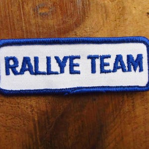 May include: A white embroidered patch with a blue border. The text "RALLYE TEAM" is embroidered in blue.