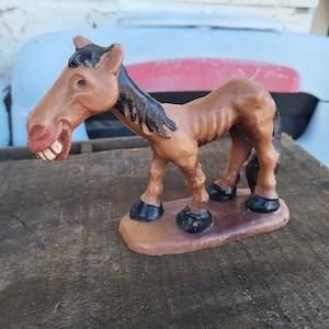 May include: A brown horse figurine with a comically exaggerated face and teeth. The horse has a black mane and tail, and black hooves. The figurine is standing on a brown base. The horse has a thin, bony appearance.