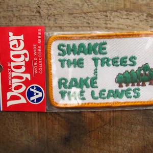 May include: A white embroidered patch with orange stitching. The patch says "Shake the Trees Rake the Leaves" with a green cartoon of a row of trees with brown trunks.
