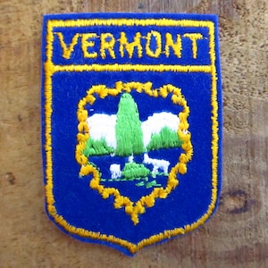 May include: A blue embroidered patch with the word "VERMONT" in yellow. The patch features a landscape scene with a green tree, white mountains, and a white deer, all outlined in yellow.