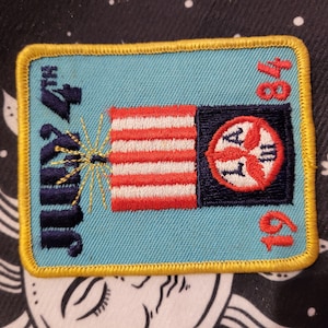 May include: A blue and yellow embroidered patch with the text "July 4th 1984" and a red, white, and blue American flag with a red, white, and blue circle with the letters "LA" in the center.