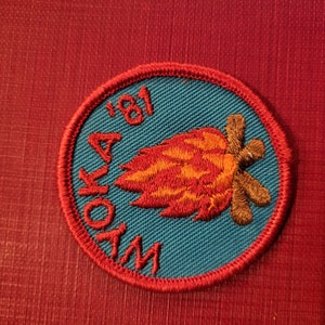 May include: A blue and red embroidered patch with the text "WOKA 181" and a stylized image of a campfire with flames and logs.