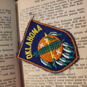 May include: A blue and orange embroidered patch with the word "OKLAHOMA" in white lettering. The patch features a stylized image of a green tree with a yellow and white spear, surrounded by white feathers.