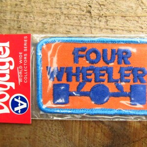 May include: A blue and orange embroidered patch with the text "Four Wheeler" and a stylized image of a four-wheeler. The patch is packaged in a clear plastic bag with a red and white Voyager label.