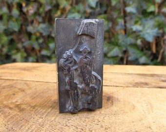 May include: A vintage metal stamp with a design of two figures on horseback, one holding a flag. The stamp is made of a dark metal and has a worn, aged appearance.
