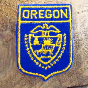 May include: A blue felt patch with yellow embroidered stitching. The patch features the state seal of Oregon, which includes a sun, a wagon, and a bird. The text "OREGON" is embroidered in yellow at the top of the patch.