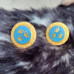 May include: A pair of gold-toned earrings with a textured border. Each earring features a light blue center with three gold crowns. The earrings are round and have a classic design.