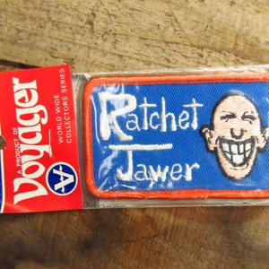 May include: A blue and orange embroidered patch with the text "Ratchet Jawer" and a cartoon image of a smiling bald man with large teeth.