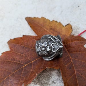 May include: A silver thimble with a floral design and a heart shape. The thimble is sitting on a brown leaf.