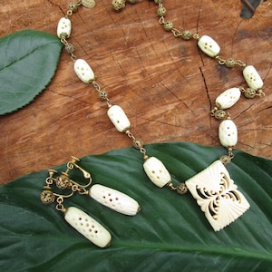 May include: An antique ivory-colored necklace and matching earrings set. The necklace features carved oval beads and a decorative pendant. The earrings have a similar design, with a clip-on style. The jewelry is set on a gold-toned chain.