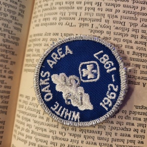 May include: A blue and white embroidered patch with the text "WHITE OAKS AREA" and a white oak leaf design. The patch also includes a white cross with a plus sign in the center. The text "1961 - 1987" is embroidered in white on the bottom right corner of the patch.