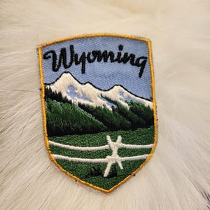 May include: A blue and green embroidered patch with the word "Wyoming" in black lettering. The patch depicts a mountain range with snow-capped peaks and a green field with a white fence.