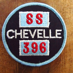 May include: A round, black patch with the words "SS CHEVELLE 396" embroidered in white and red. The patch has a light blue border and is set against a wooden background.