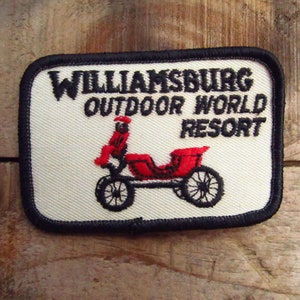May include: White embroidered patch with black stitching. The patch features the text "Williamsburg Outdoor World Resort" and a black and red illustration of a horse-drawn carriage.