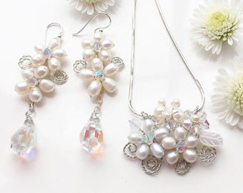 May include: A set of delicate pearl and crystal jewelry featuring a necklace and earrings. The necklace and earrings are made with white pearls and iridescent crystals, and are arranged in a floral design. The jewelry is perfect for a wedding or other special occasion.