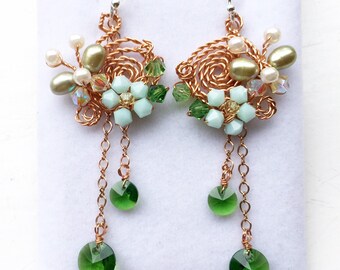May include: A pair of handcrafted earrings featuring intricate copper wirework. The earrings showcase a floral design with light green and mint-colored beads, along with dangling green crystal accents. Small white pearls and iridescent crystals add sparkle.